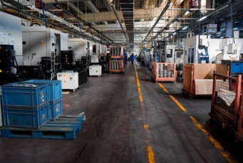 Blick durch eine industrielle Fertigungshalle mit einer Reihe von CNC-Maschinen und Materialtransportbehältern entlang des Gangs